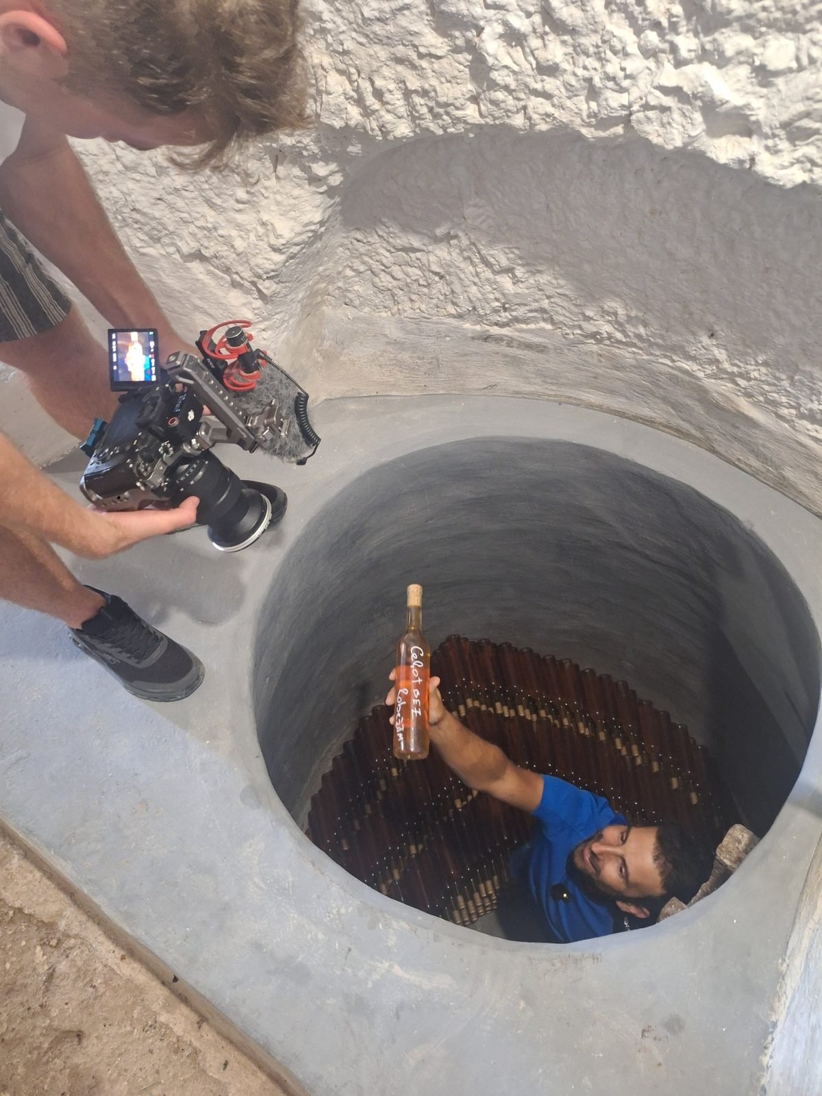 A host hands up a bottle labelled Ceļot Bez Robežām from a round underground cellar while the crew films, September 2026