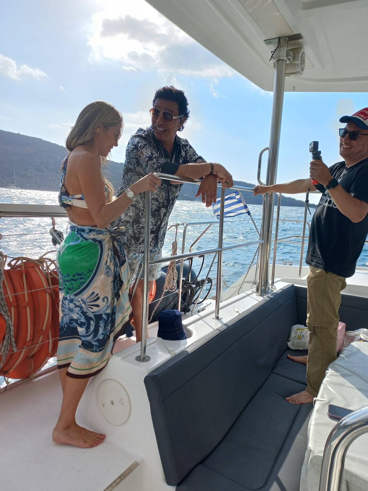 The presenters on the catamaran deck while a crew member films, La Vuelta al Mundo en 80 Risas, Santorini, October 2022